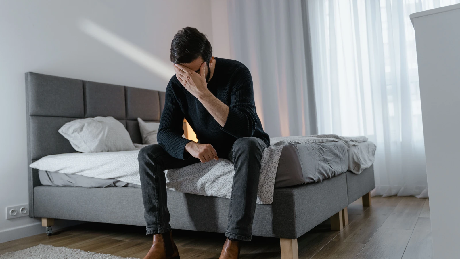 Un uomo pensieroso in un maglione nero è seduto su un letto in una camera da letto moderna.
