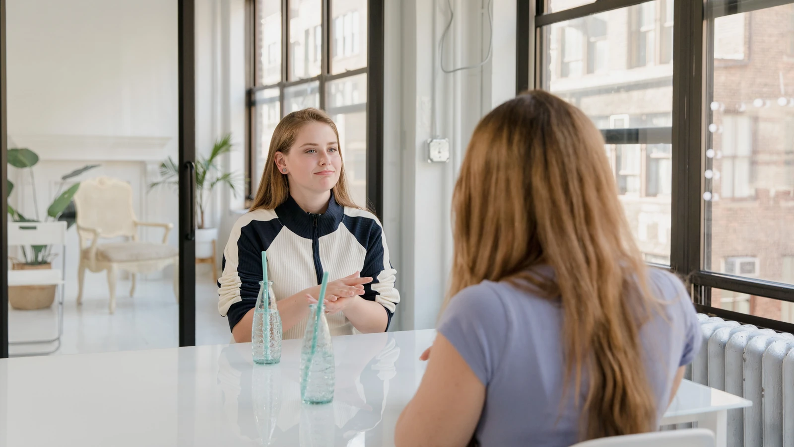 Due donne conversano in un interno moderno con grandi finestre e luce naturale.