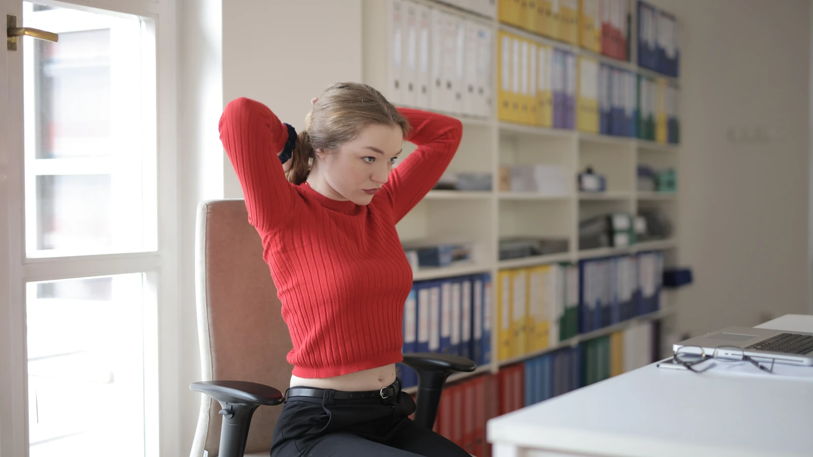 Una giovane donna in un maglione rosso si allunga alla sua scrivania in un moderno spazio ufficio.