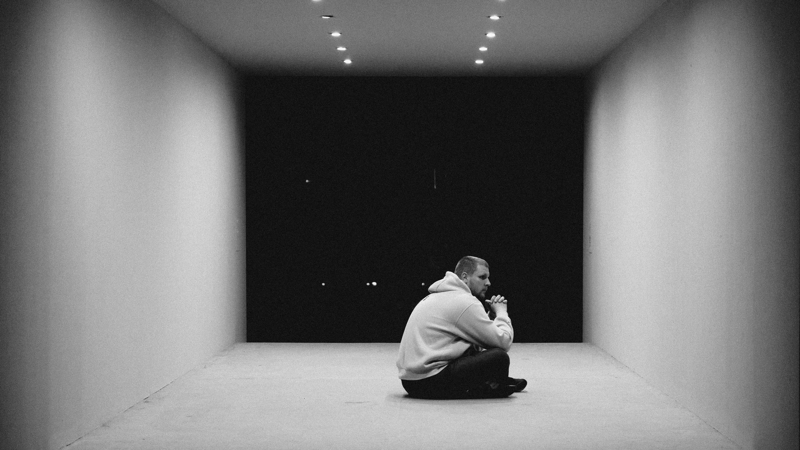 Un uomo è seduto da solo in uno spazio minimalistico e monocromatico, riflettendo un senso di solitudine.