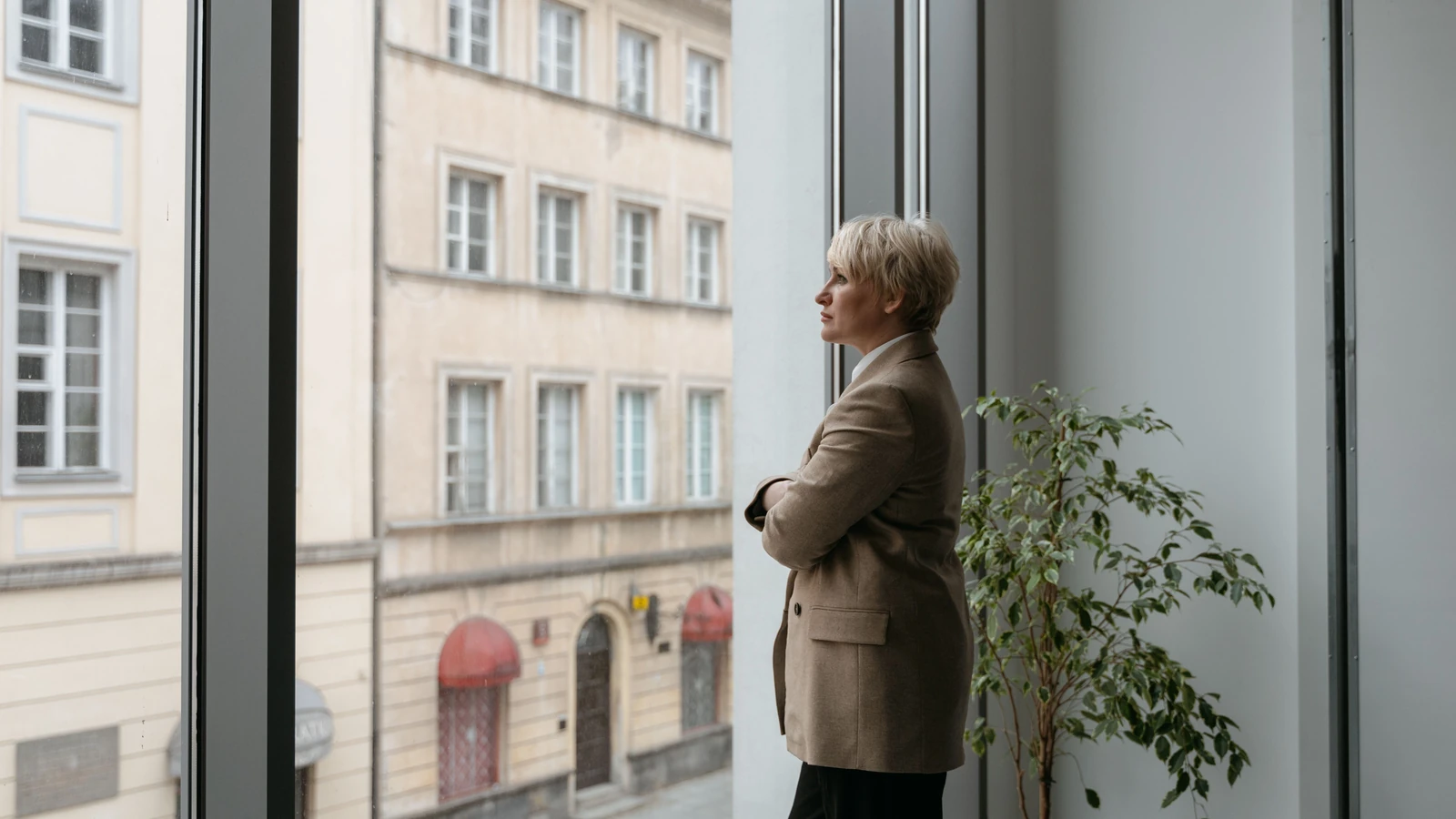 Una donna pensierosa è accanto a una grande finestra a guardare il panorama cittadino all'esterno, con le braccia incrociate.