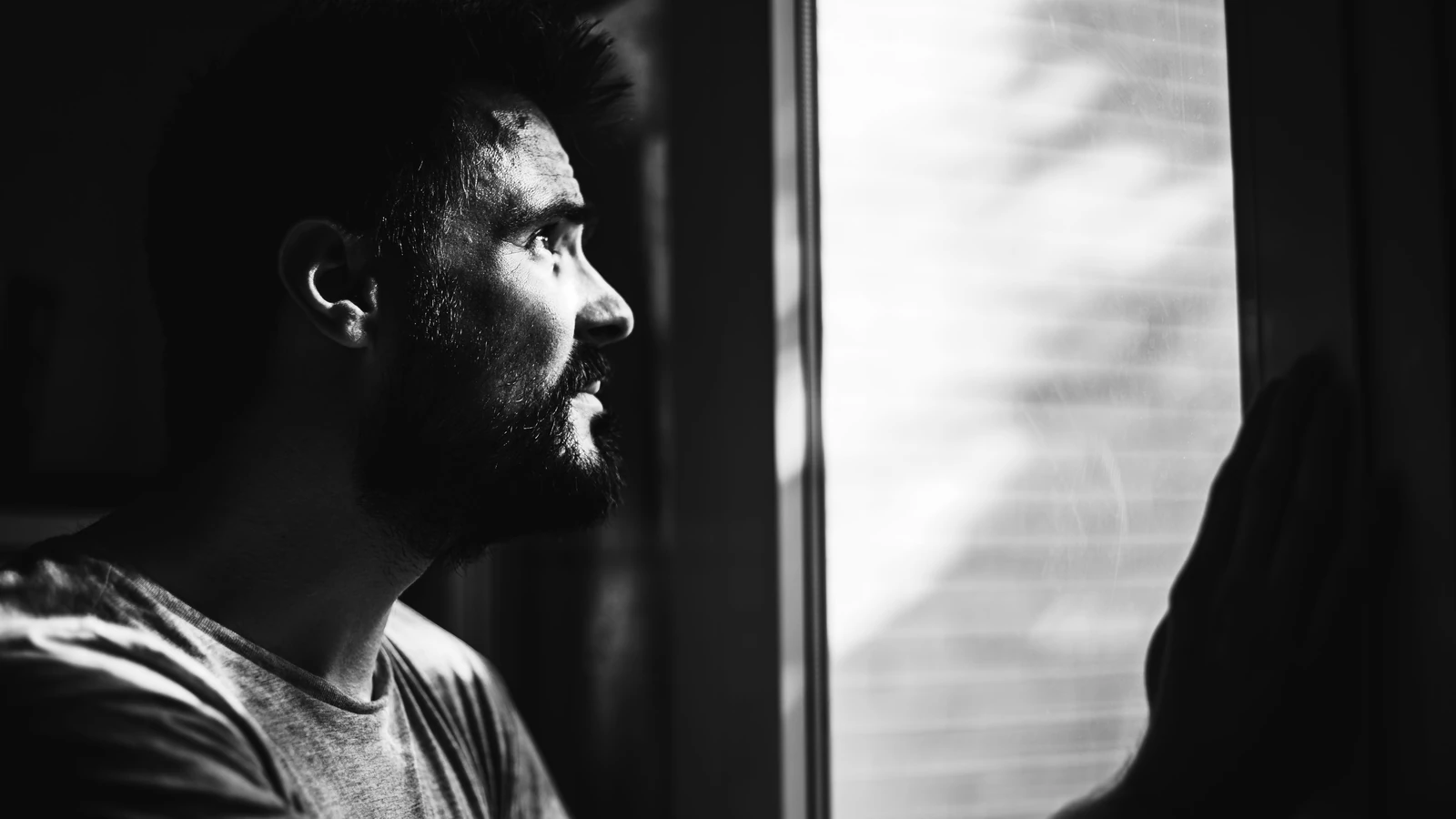 Un uomo riflessivo con barba guarda fuori da una finestra, catturato in drammatico bianco e nero.