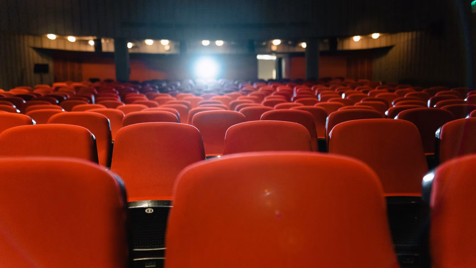 Una vista di un cinema vuoto con file di sedili rossi rivolti verso lo schermo illuminato. — Tima Miroshnichenko – Pexels