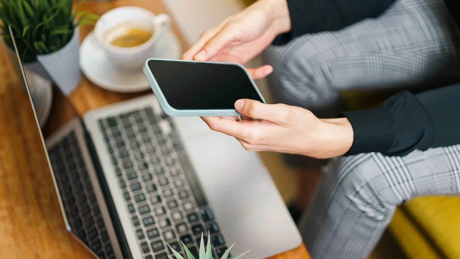 Primo piano di mani che tengono uno smartphone sopra un laptop, con caffè e piante su una scrivania in legno.