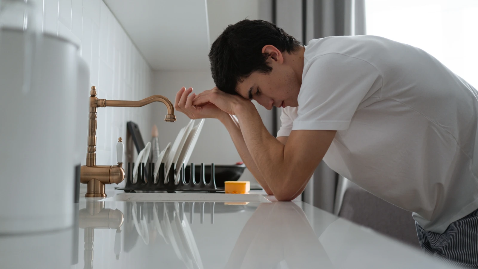 Un giovane appare stanco e stressato, appoggiato a un piano di cucina in contemplazione.