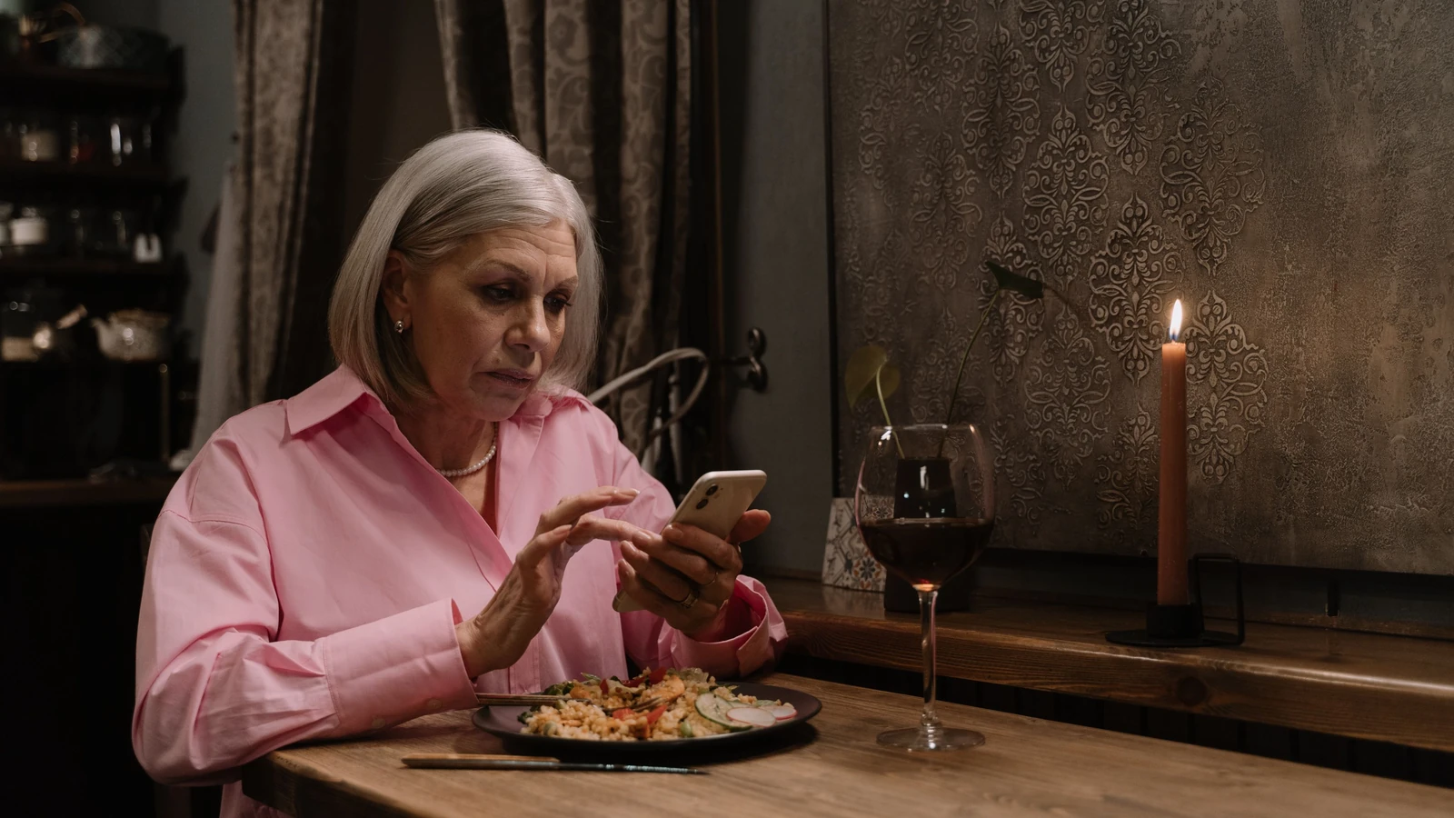 Donna anziana che cena da sola usando uno smartphone a lume di candela con vino rosso all'interno.