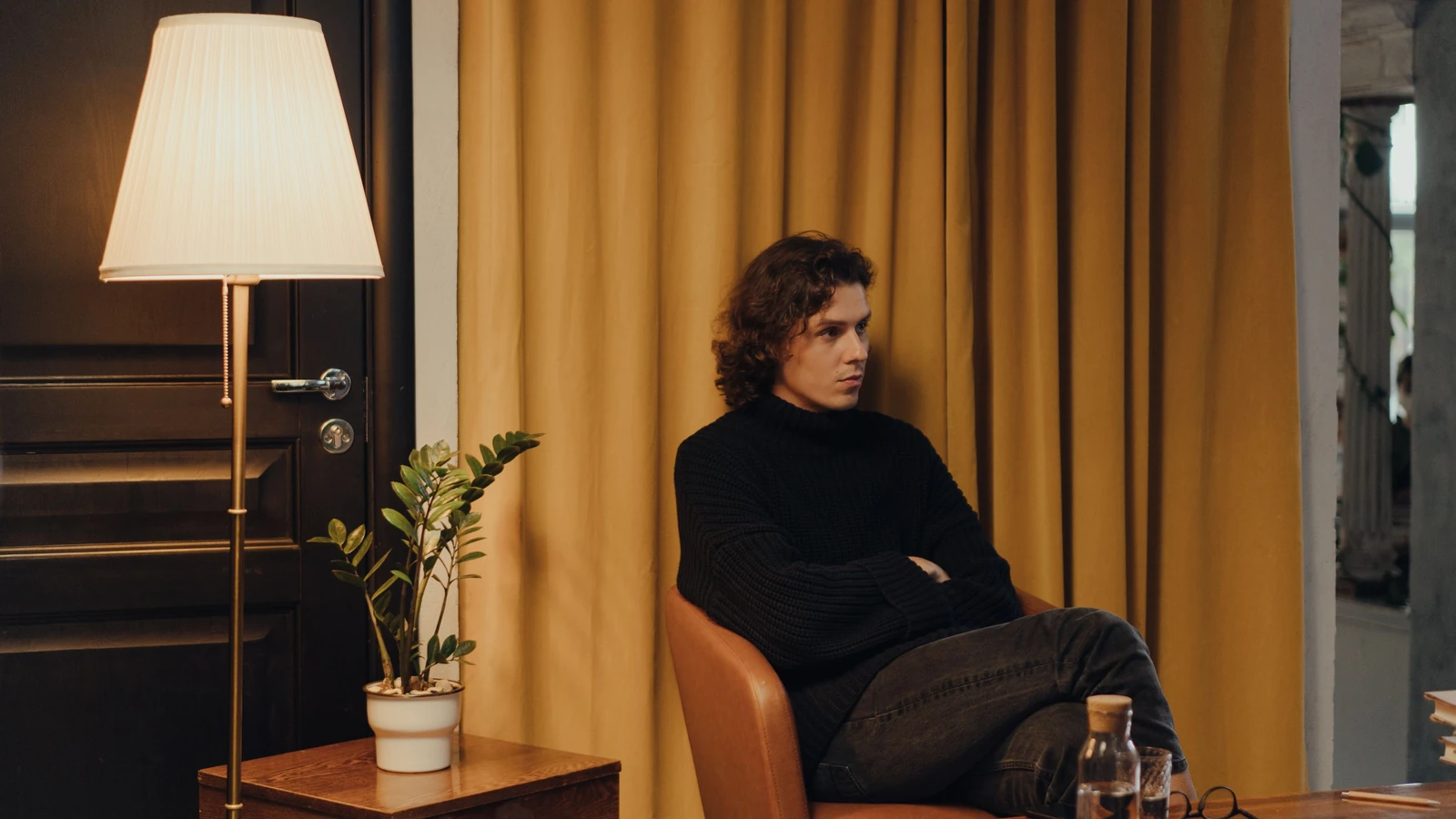 Uomo in maglione nero seduto all'interno accanto a una tenda gialla e a una lampada da terra.