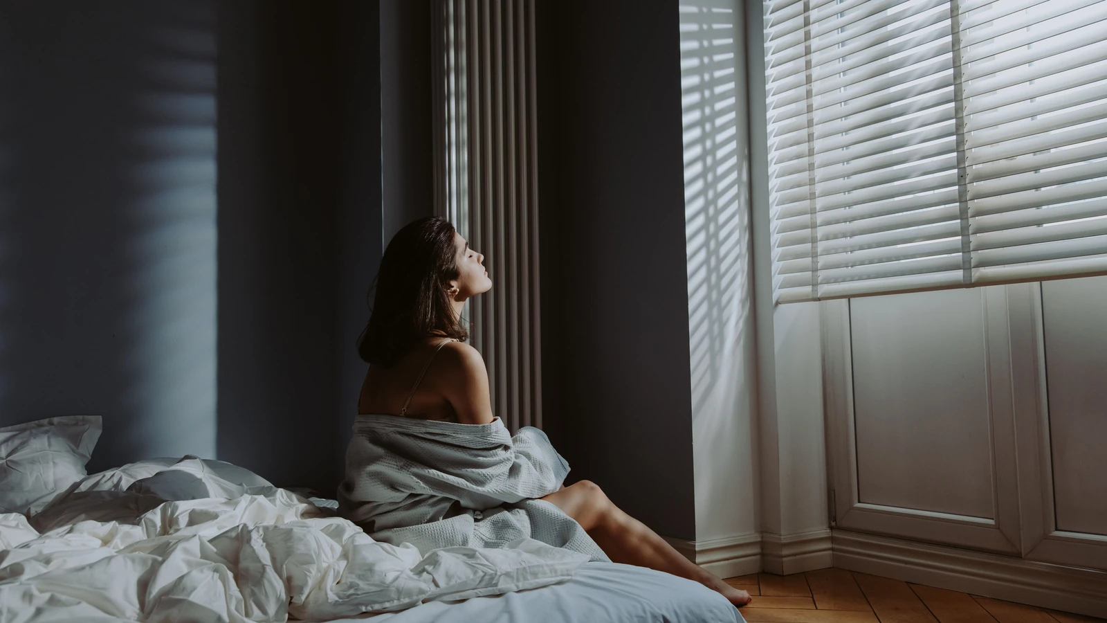 Donna seduta sul letto in una camera da letto accogliente, avvolta in una coperta con la luce del sole che entra dalle veneziane. — cottonbro studio – Pexels
