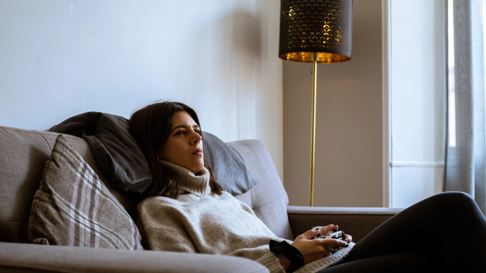 Donna in maglione a collo alto che si rilassa con un controller di gioco su un divano accogliente nel soggiorno di Edimburgo.