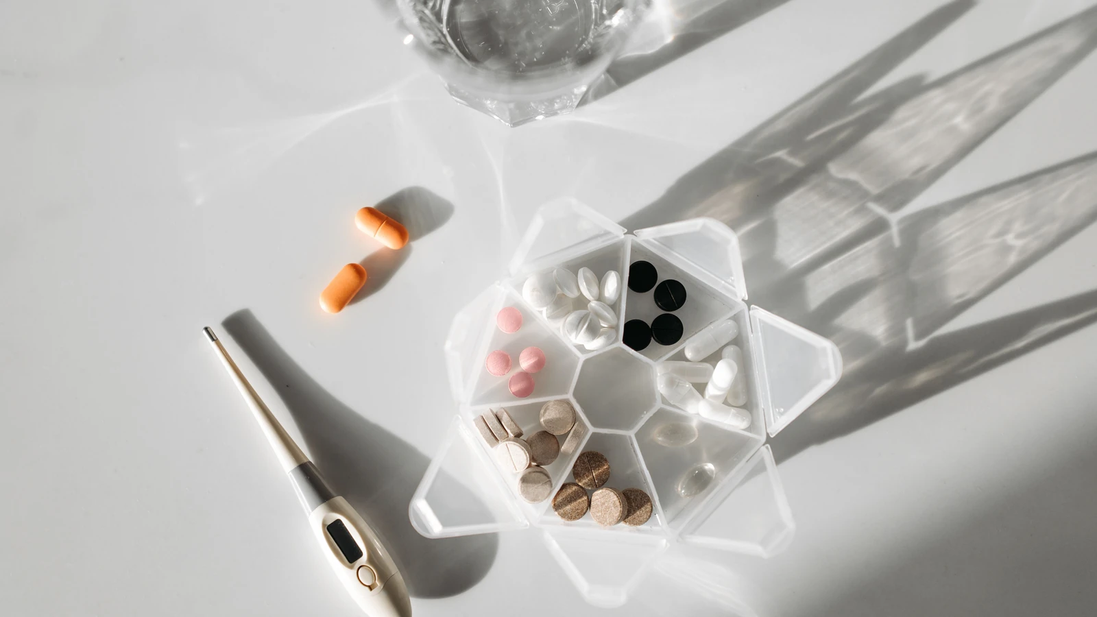 Un'immagine dettagliata che mostra un organizzatore di pillole, un termometro digitale e pillole con un bicchiere d'acqua su una superficie bianca. — Pavel Danilyuk – Pexels