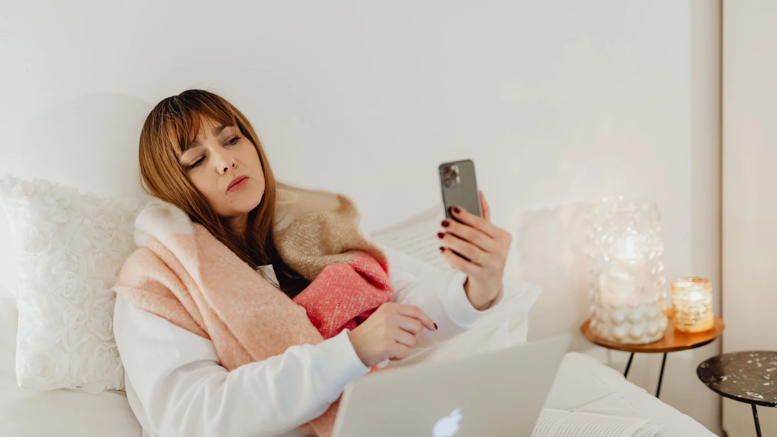 Una donna avvolta in una sciarpa che utilizza il suo smartphone e laptop in un ambiente accogliente di camera da letto.