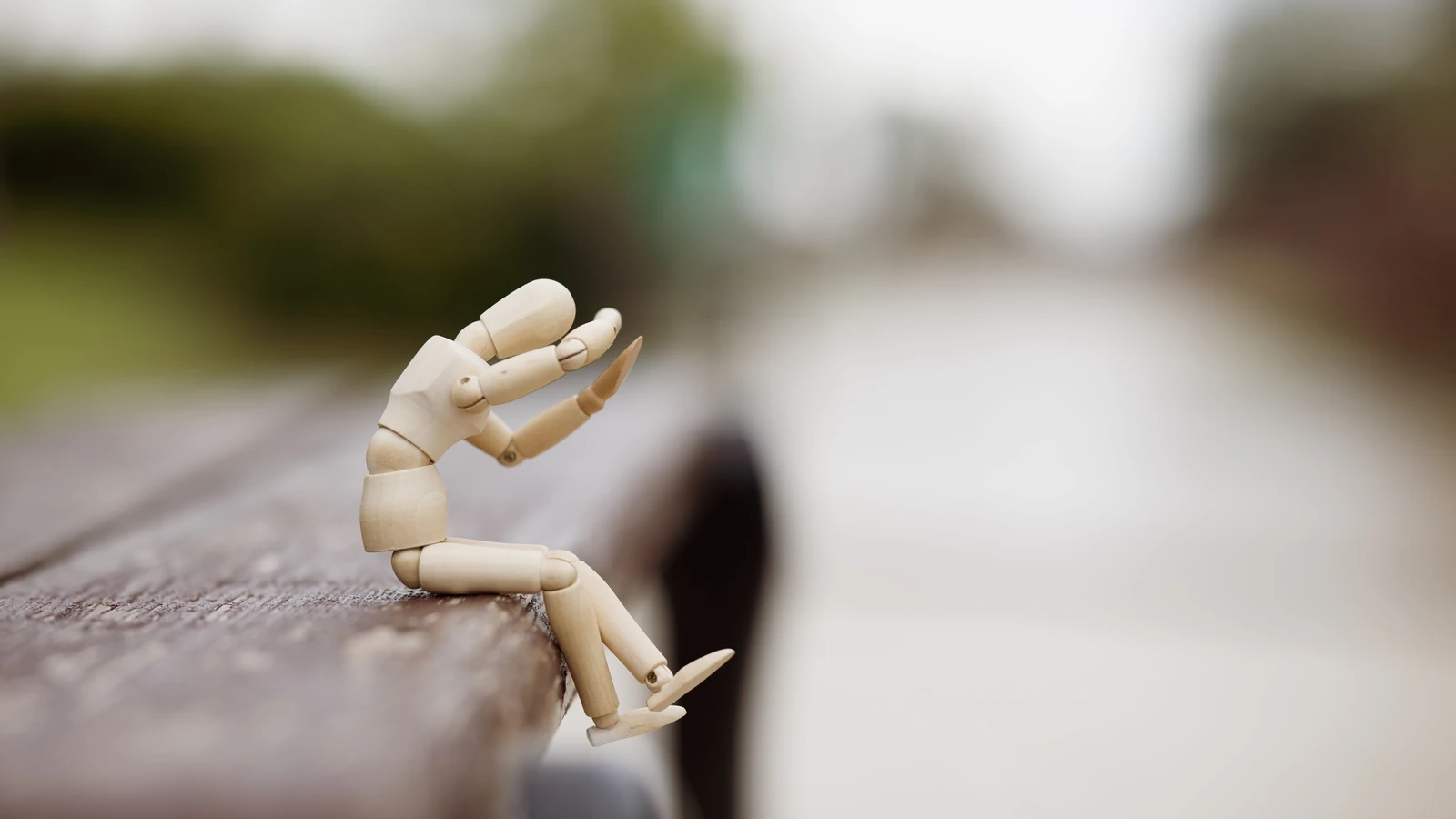 Figurina manichino di legno seduta da sola su una panchina all'aperto, esprimendo solitudine e contemplazione.