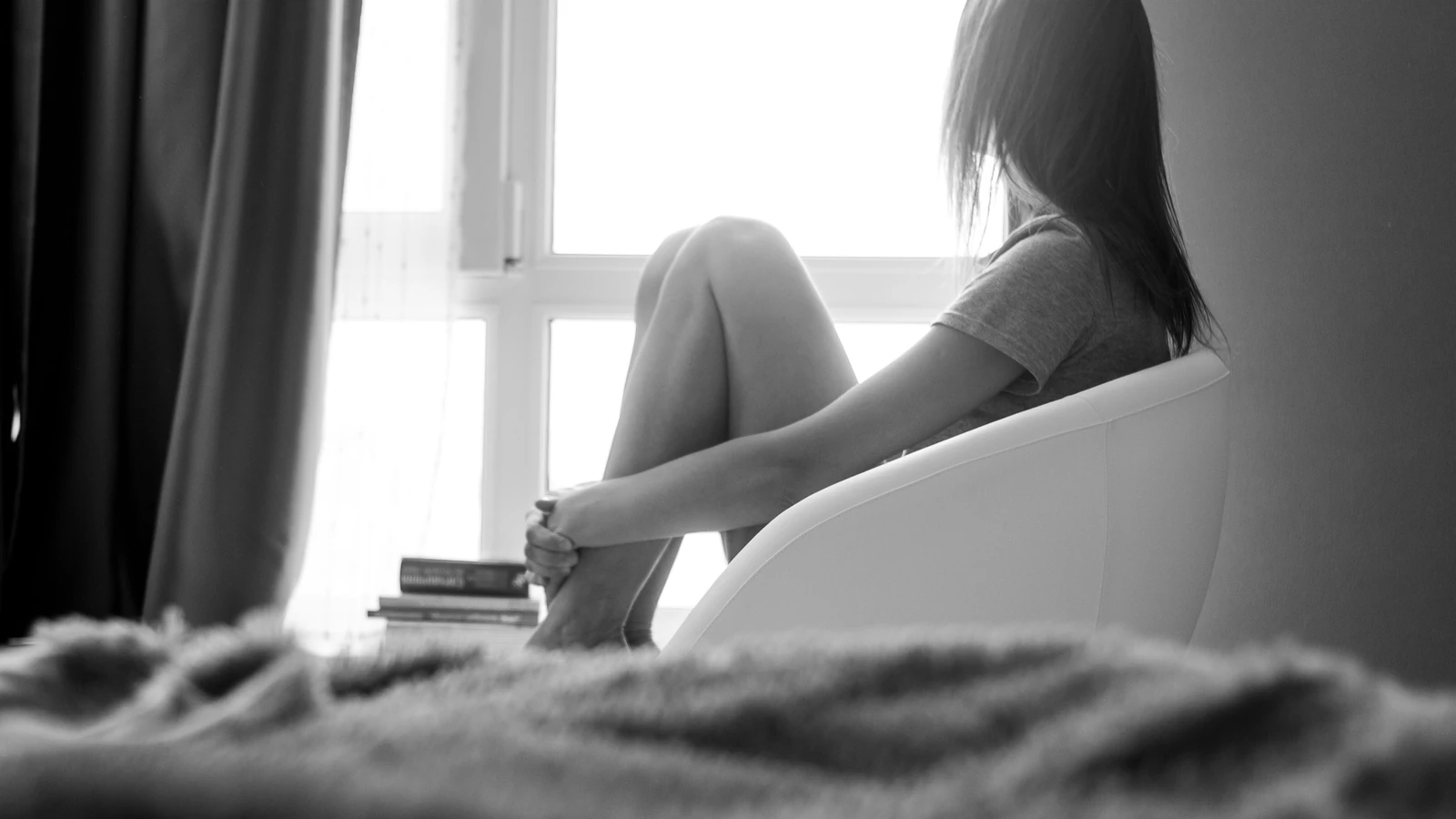 Una donna pensierosa è seduta su una sedia vicino a una finestra luminosa in un ambiente monocromatico.