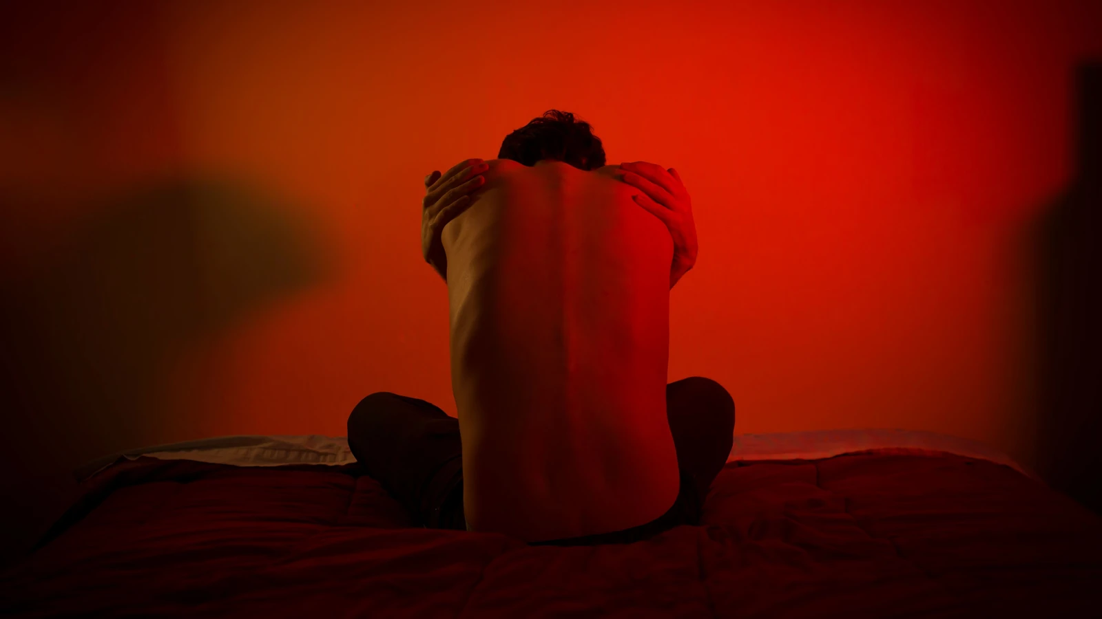 Un uomo seduto solo su un letto in una stanza illuminata di rosso, che rappresenta solitudine e introspezione.