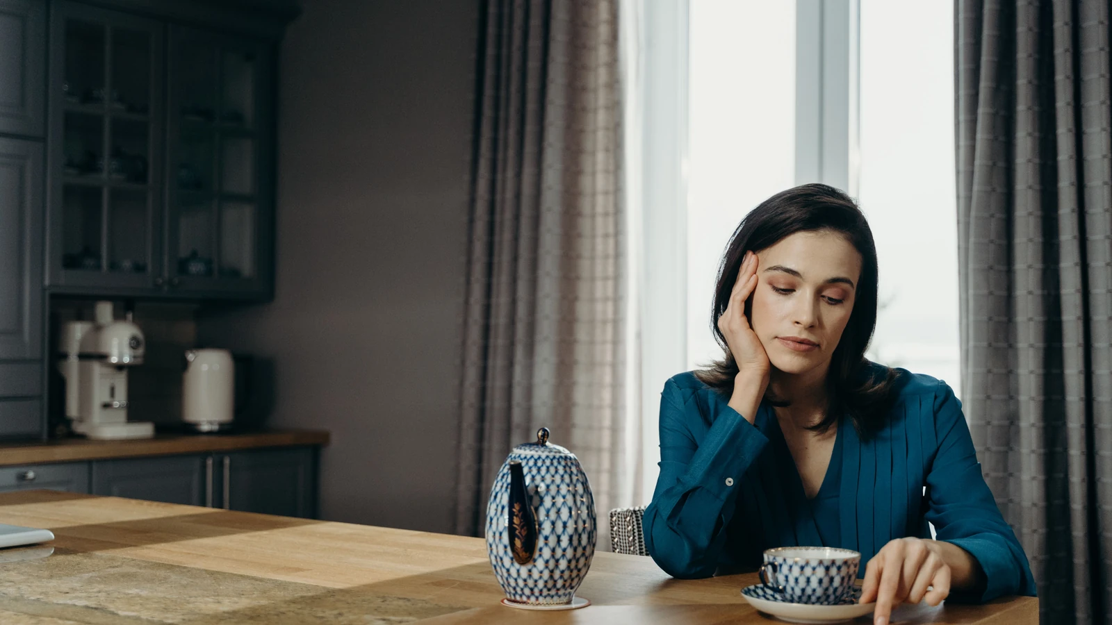Donna con una blusa blu che si rilassa con un caffè a un tavolo di legno in una sala da pranzo accogliente.