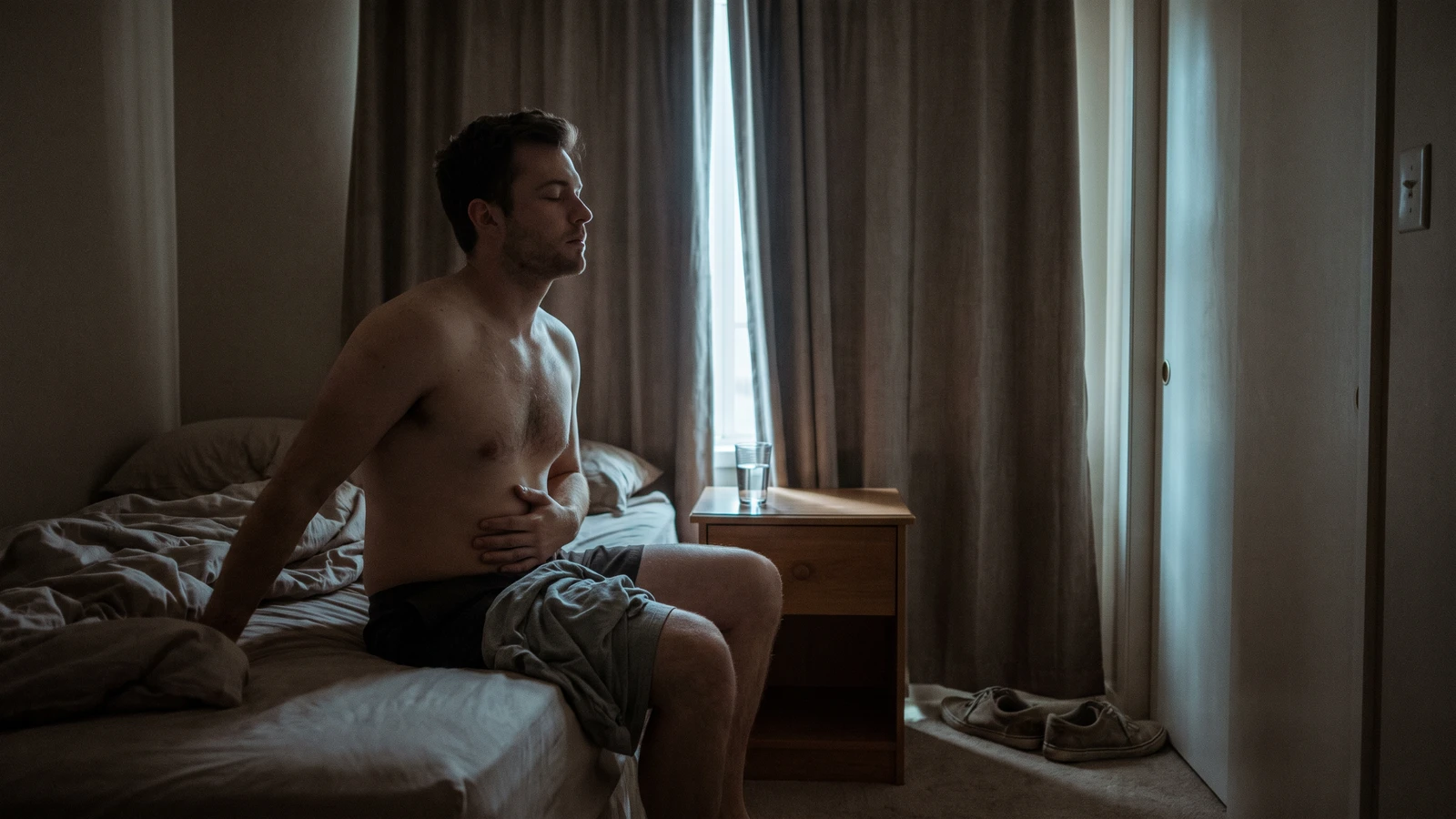 Uomo seduto sul bordo del letto al mattino con una mano sull'addome e gli occhi chiusi