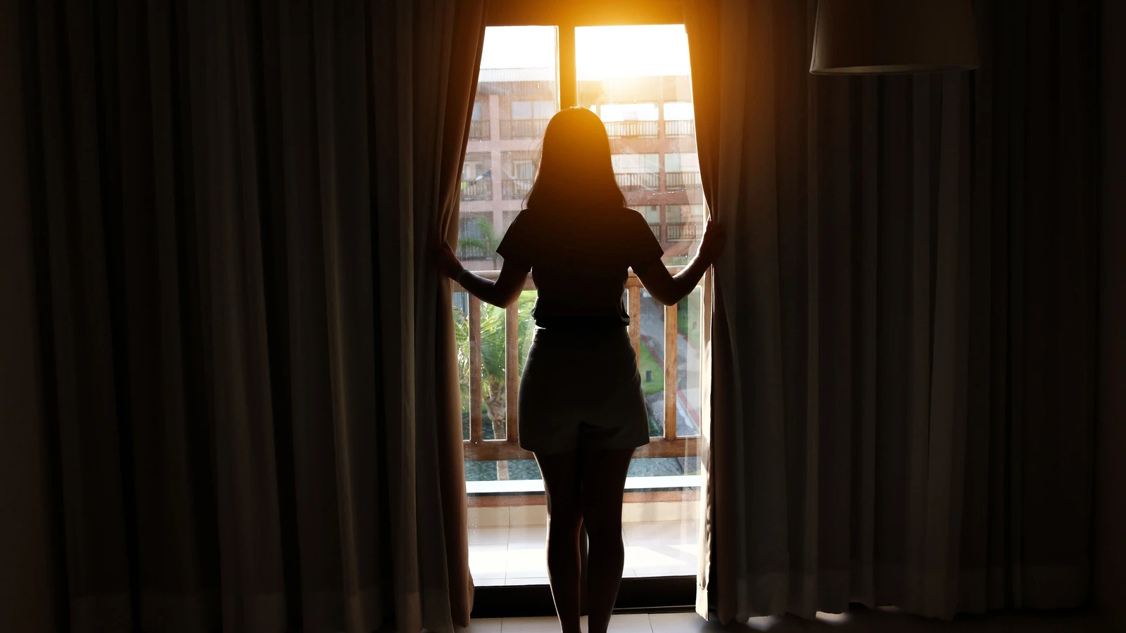 Silhouette di una donna che apre le tende all'alba in una stanza, catturando una scena mattutina tranquilla.