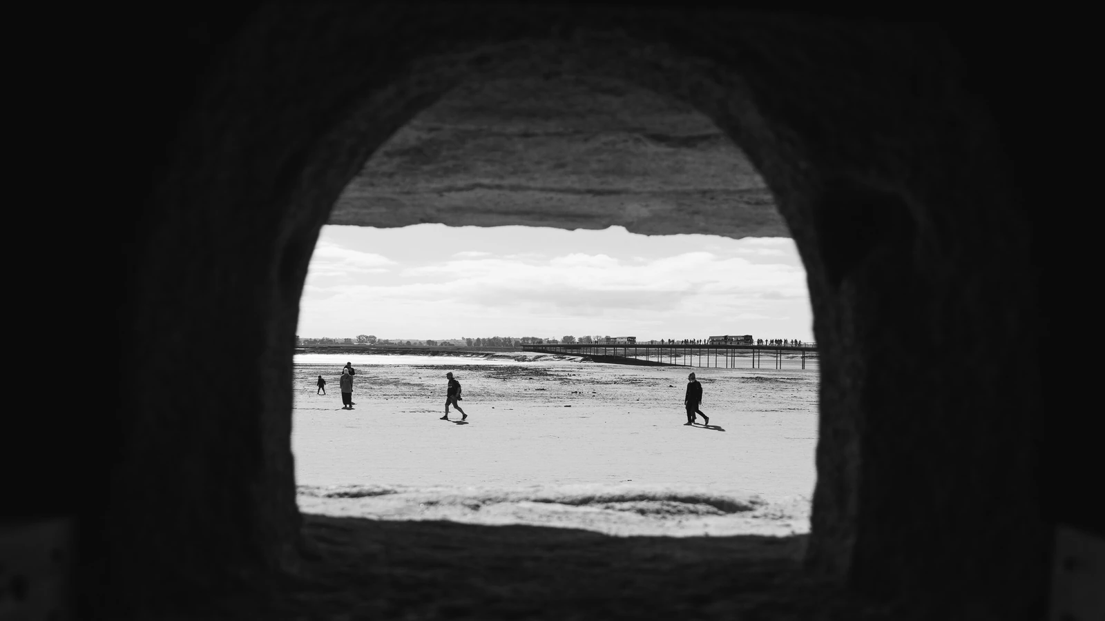 Vista in bianco e nero di persone che camminano su una spiaggia attraverso un arco di pietra, catturando silhouette serene a Mont Saint-Michel.