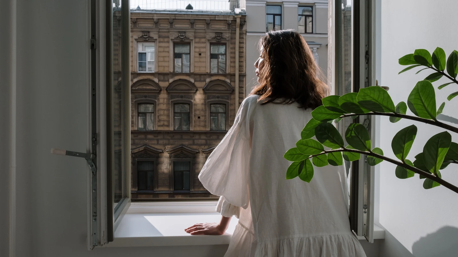 Donna in abito bianco che guarda da una finestra aperta verso un'architettura classica in una giornata soleggiata.
