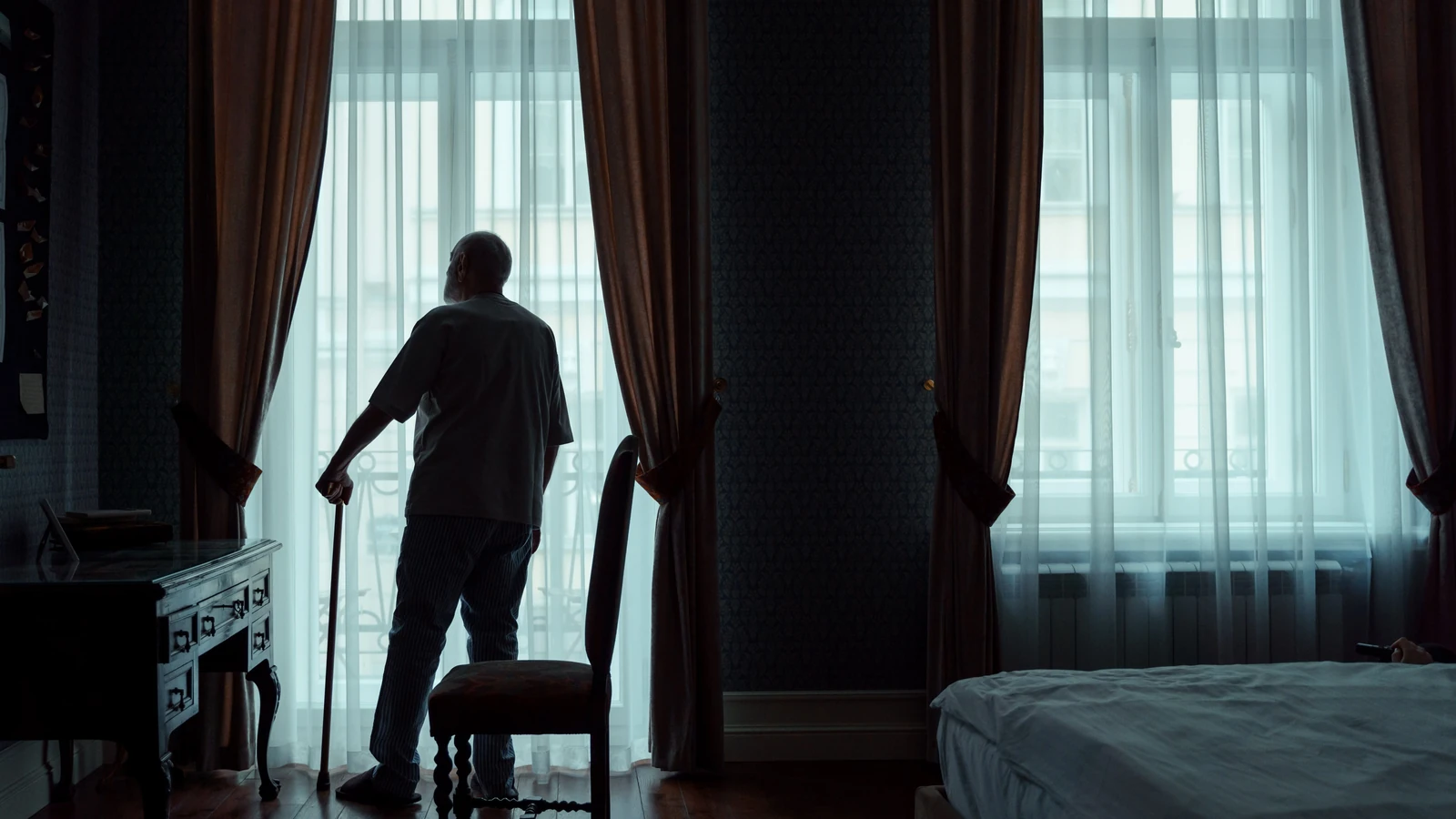 Silhouette di un uomo anziano in piedi con un bastone, che guarda fuori da una finestra coperta da una tenda.