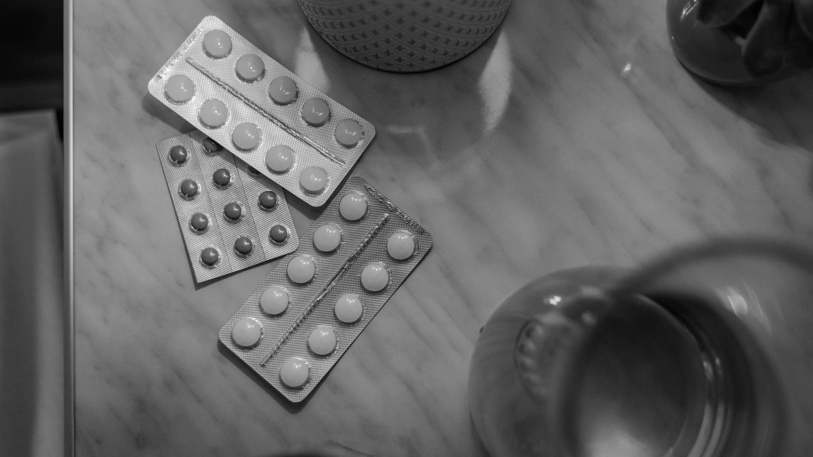 Immagine in bianco e nero che mostra blister di medicinali su un tavolo con illuminazione soffusa. — Tima Miroshnichenko – Pexels
