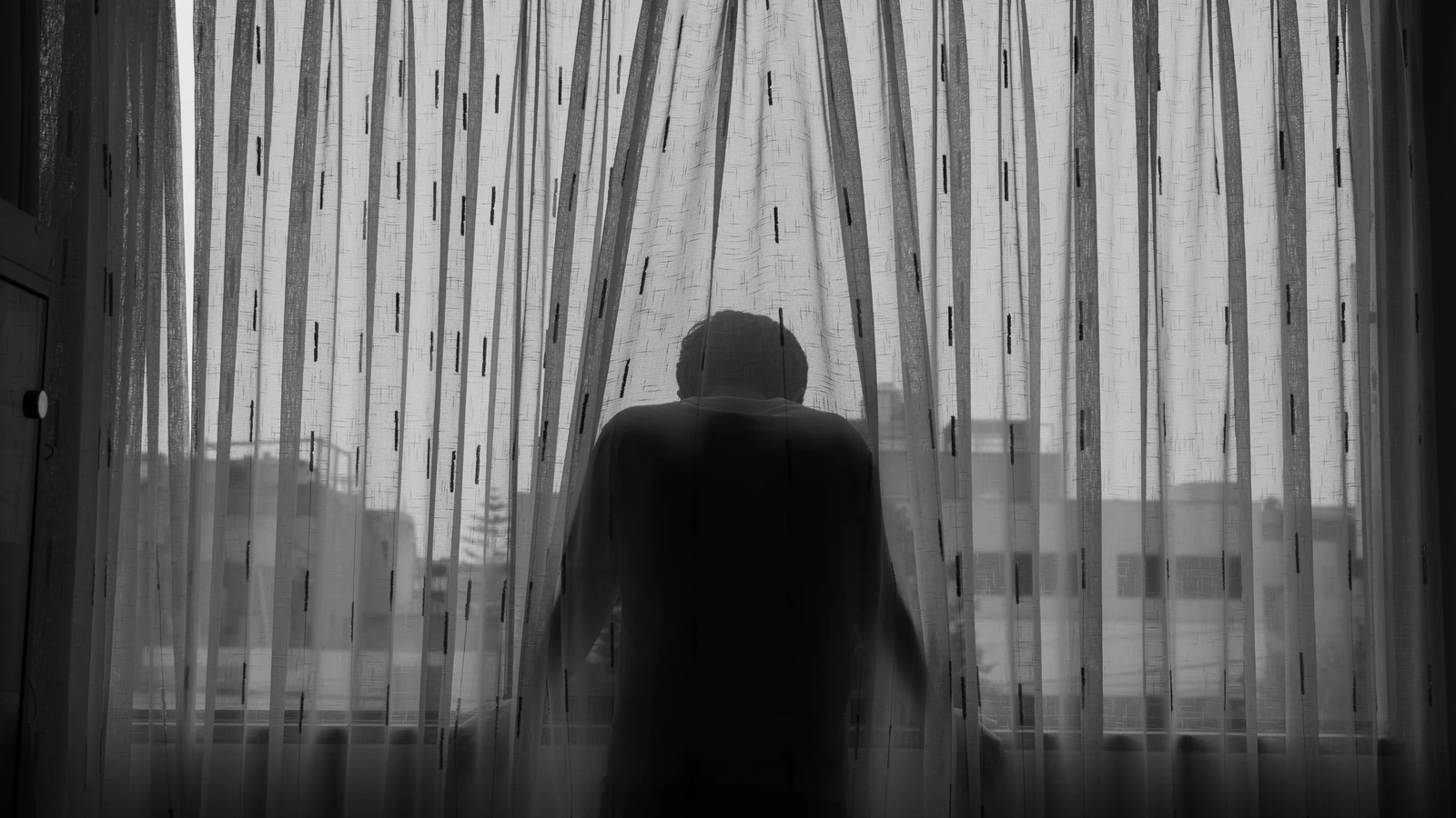Foto in bianco e nero di una persona in piedi dietro una tenda che guarda dalla finestra, trasmettendo isolamento