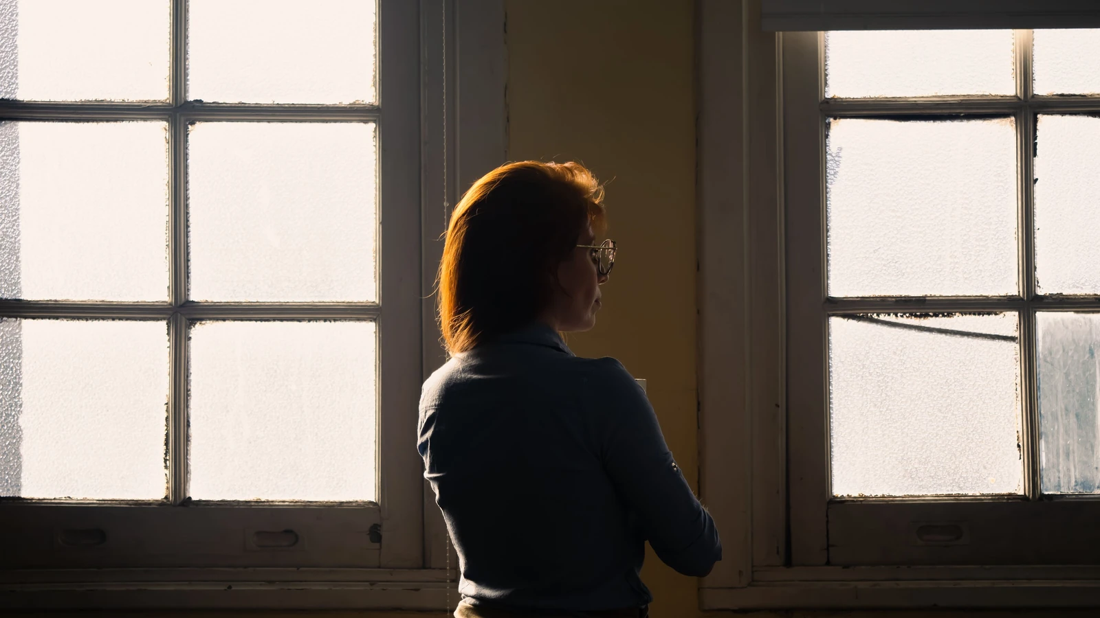Una donna pensierosa si staglia in controluce contro una finestra antica e satinata, evocando contemplazione e solitudine.