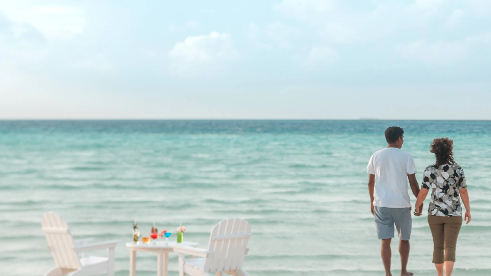 Coppia che gode di una passeggiata serena su una spiaggia tropicale con sedie e bevande.