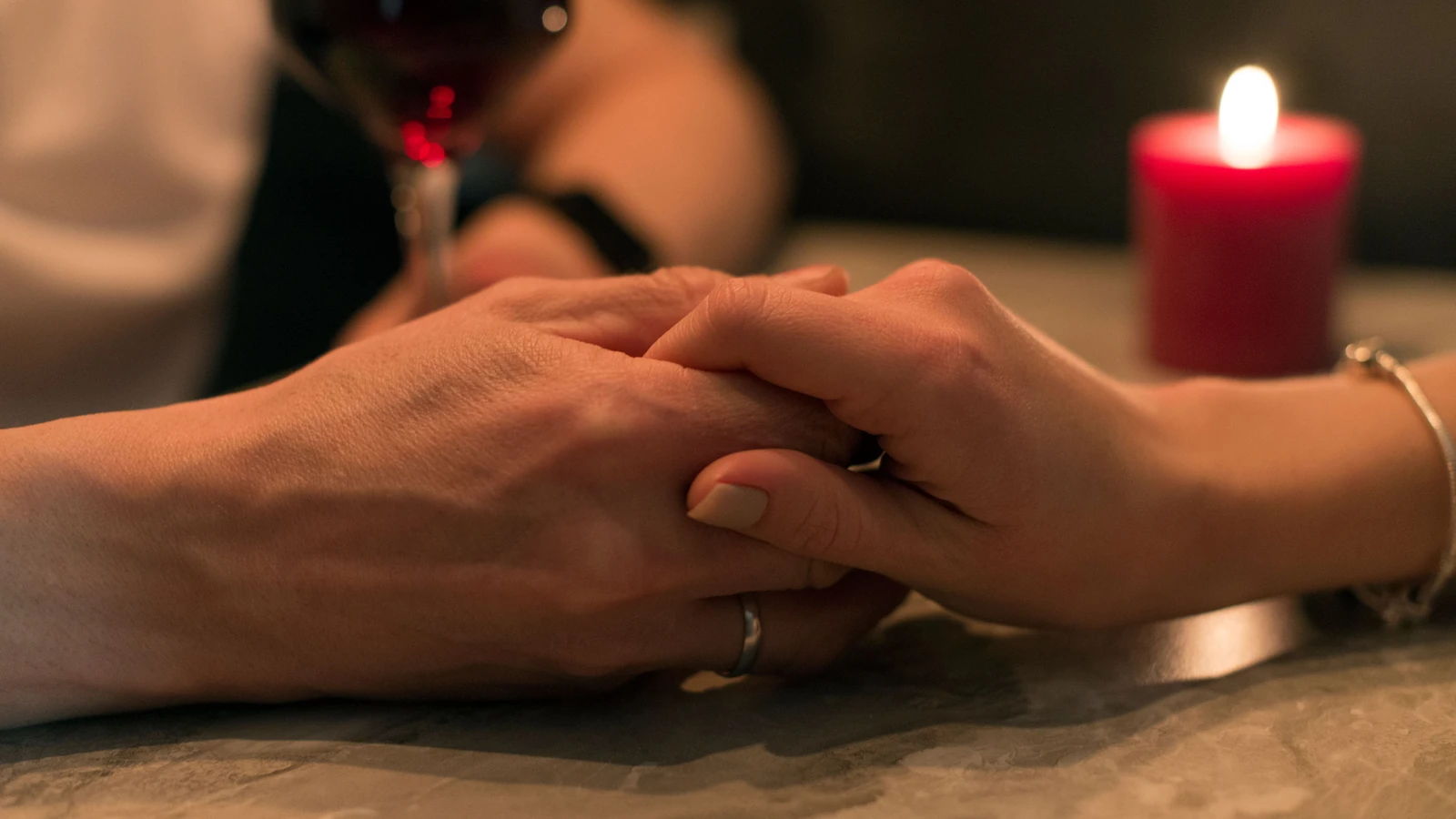 Una coppia romantica che si tiene per mano con una candela e un bicchiere di vino in una luce soffusa, simbolizzando amore e intimità.