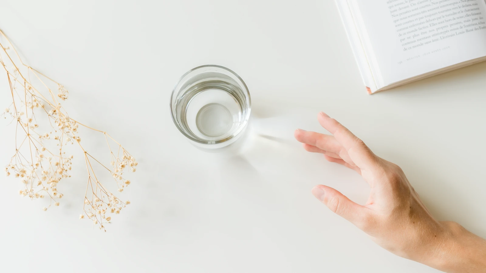 Una composizione minimalista con una mano che si allunga verso un bicchiere d'acqua su una superficie bianca accanto a un libro e fiori secchi.