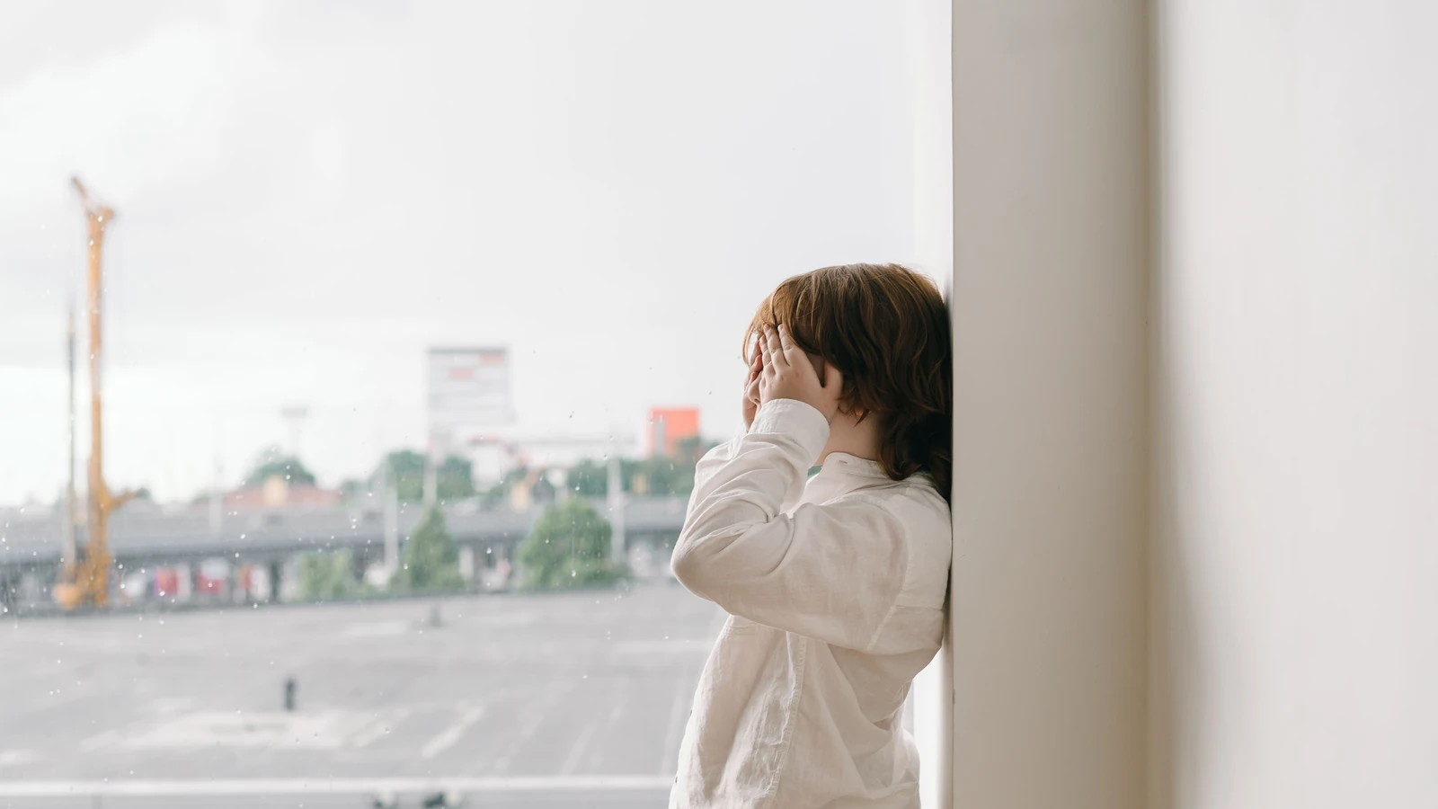 Un bambino con il volto coperto guarda fuori da una finestra su una città.
