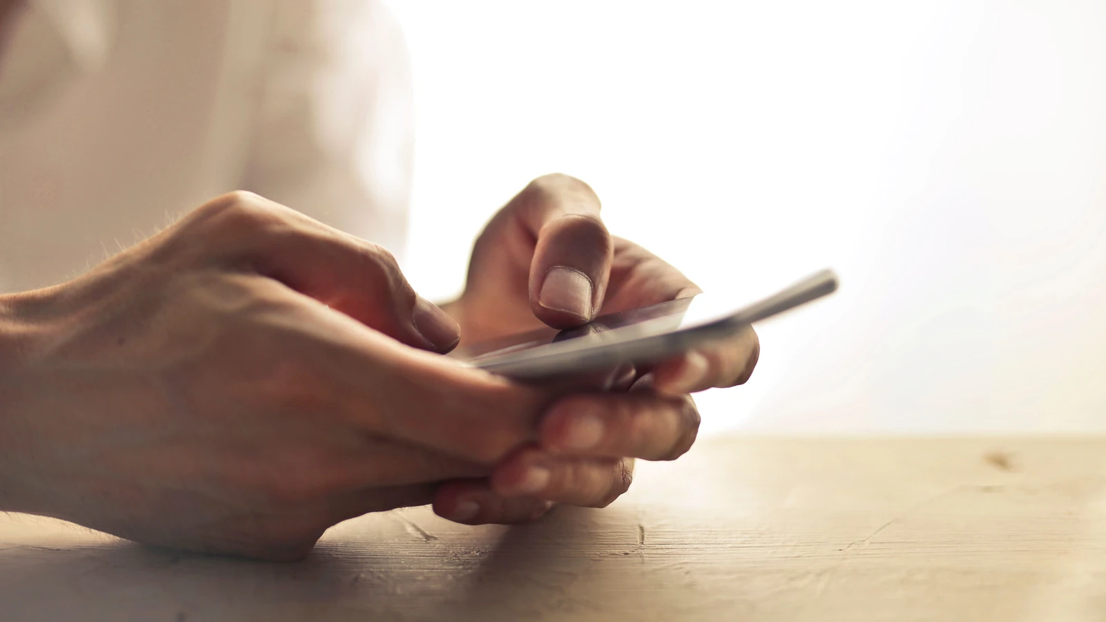Primo piano delle mani di una persona che tiene uno smartphone su un tavolo al chiaro.