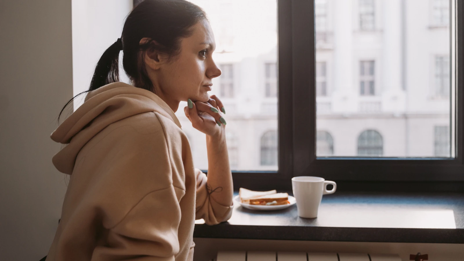 Vista laterale di una donna in felpa seduta vicino a una finestra, che contempla mentre guarda fuori. — MART  PRODUCTION – Pexels