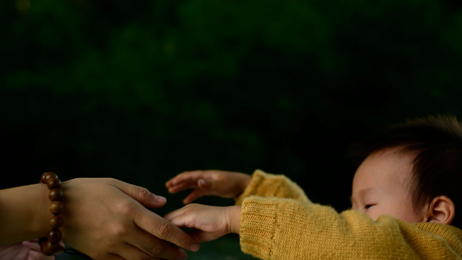 Primo piano di un bebé che si allunga per tenere la mano di un caregiver su uno sfondo verde scuro.
