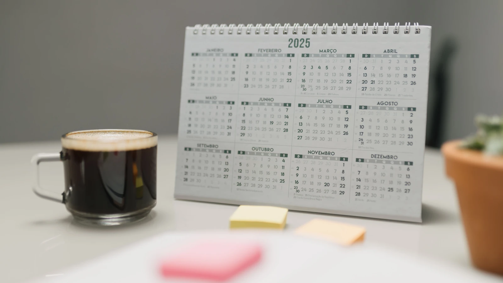 Un calendario del 2025 su una scrivania con caffè, post-it e una pianta in un ambiente d'ufficio.