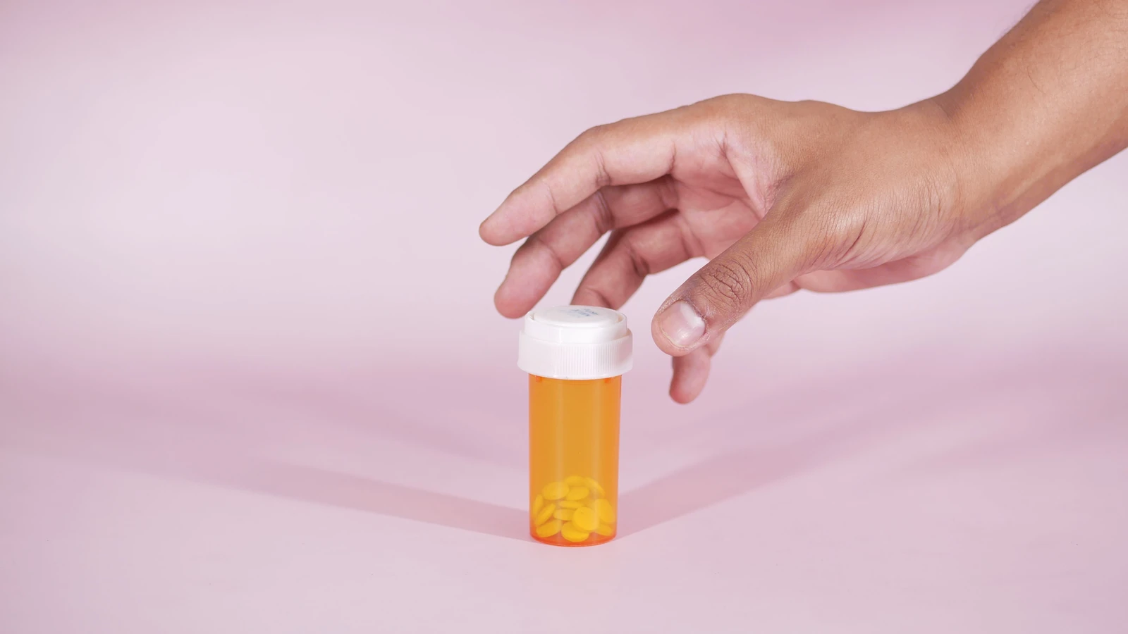 Una mano raggiunge una bottiglia di prescrizione con pillole gialle su uno sfondo rosa, simboleggiando la sanità.