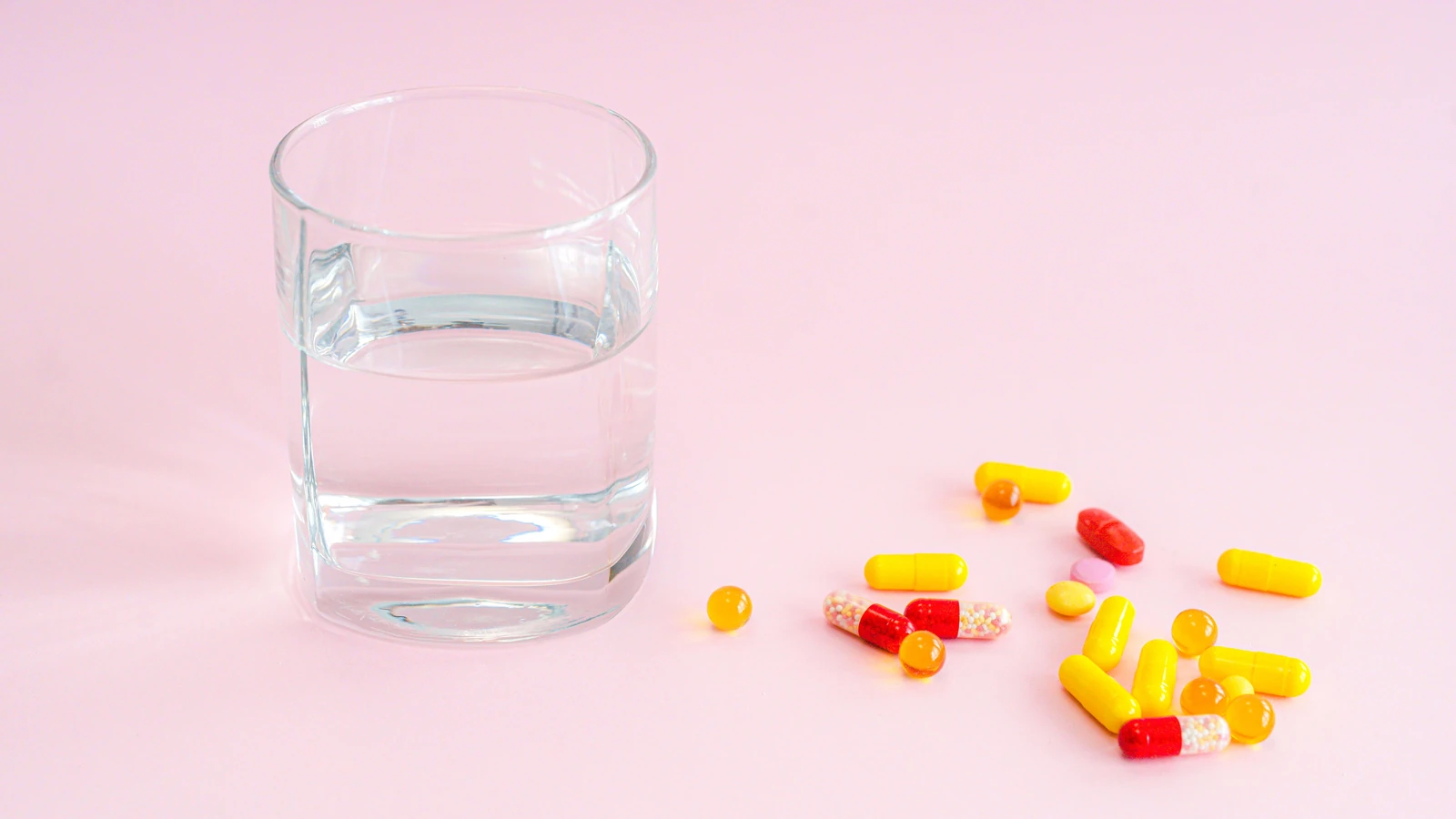 Un bicchiere d'acqua e una varietà di pillole su uno sfondo rosa, che simboleggia la medicazione. — SHVETS production – Pexels