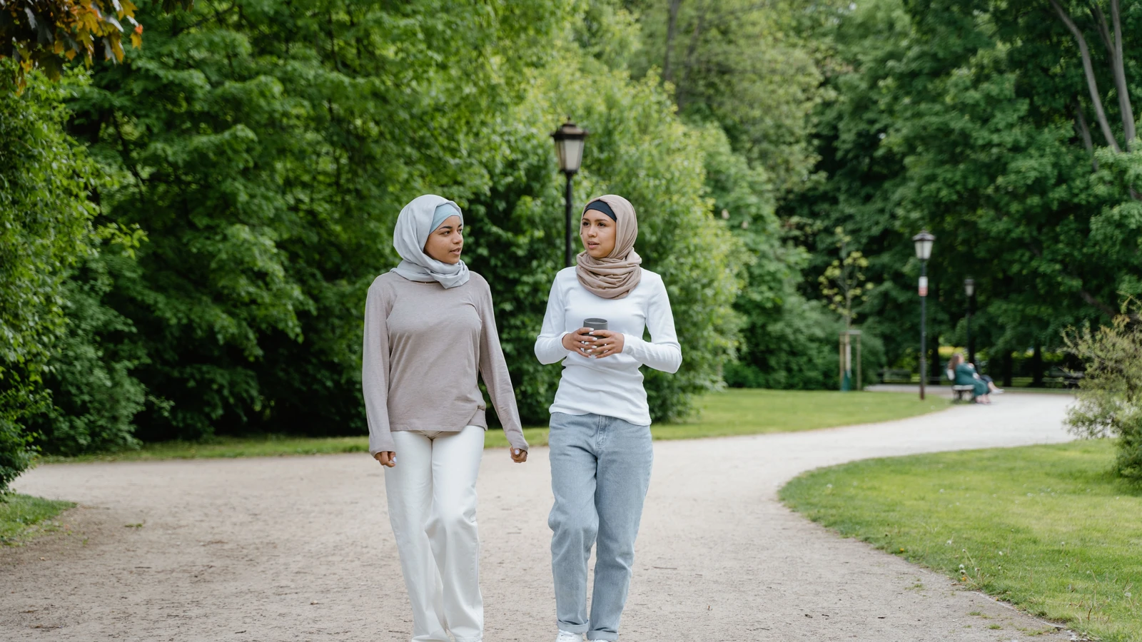 Due donne in hijab che camminano e conversano in un parco verdeggiante in una giornata di sole.