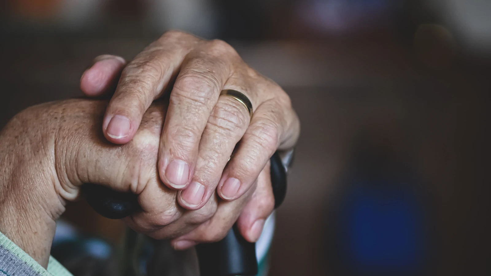 Primo piano di mani anziane che si stringono attorno a un bastone, evidenziando una fede nuziale, simbolo di amore e supporto.