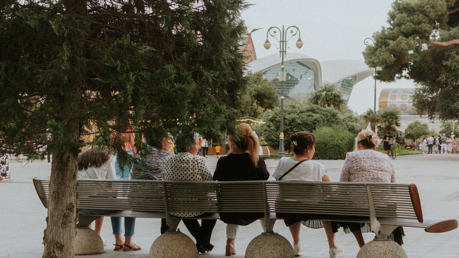 Gruppo di adulti e bambini seduti su una panchina in un parco urbano con architettura moderna.