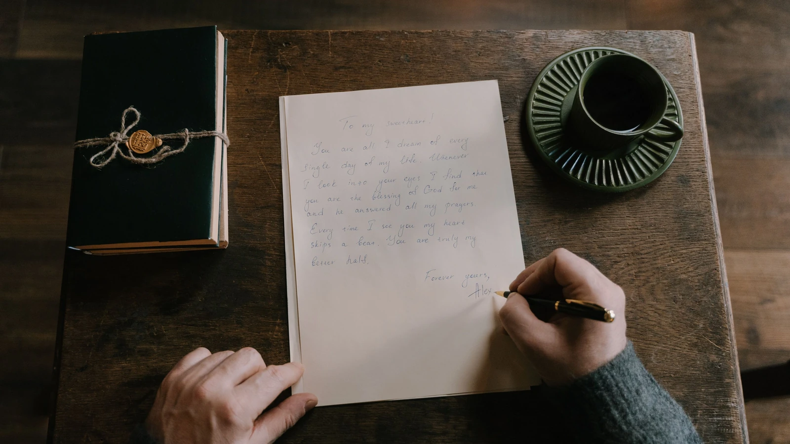 Una persona scrive a mano una lettera accanto a un diario e una tazza di caffè su un tavolo di legno.