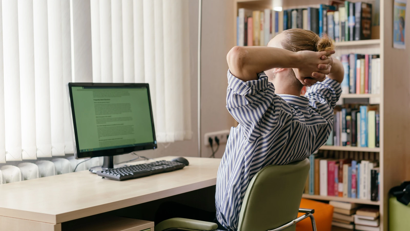 Un giovane adulto è seduto a una scrivania, mani sulla testa, in uno spazio di studio con un computer e scaffali di libri.