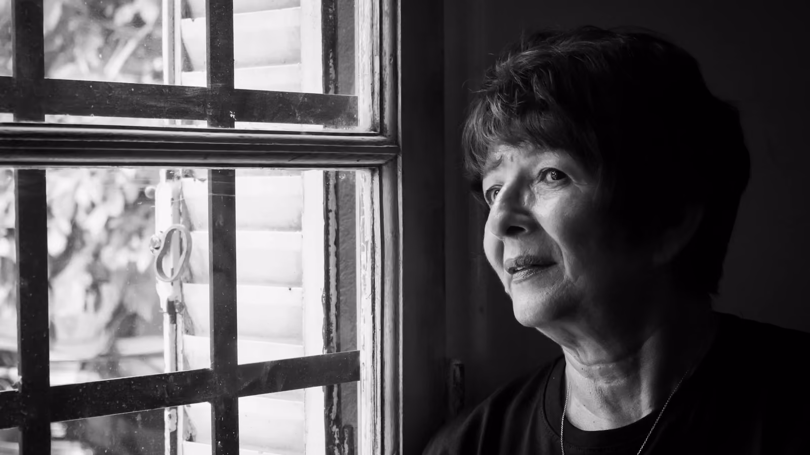 Ritratto in bianco e nero di un'anziana signora che guarda pensierosa fuori da una finestra in casa. — Kari Alfonso – Pexels
