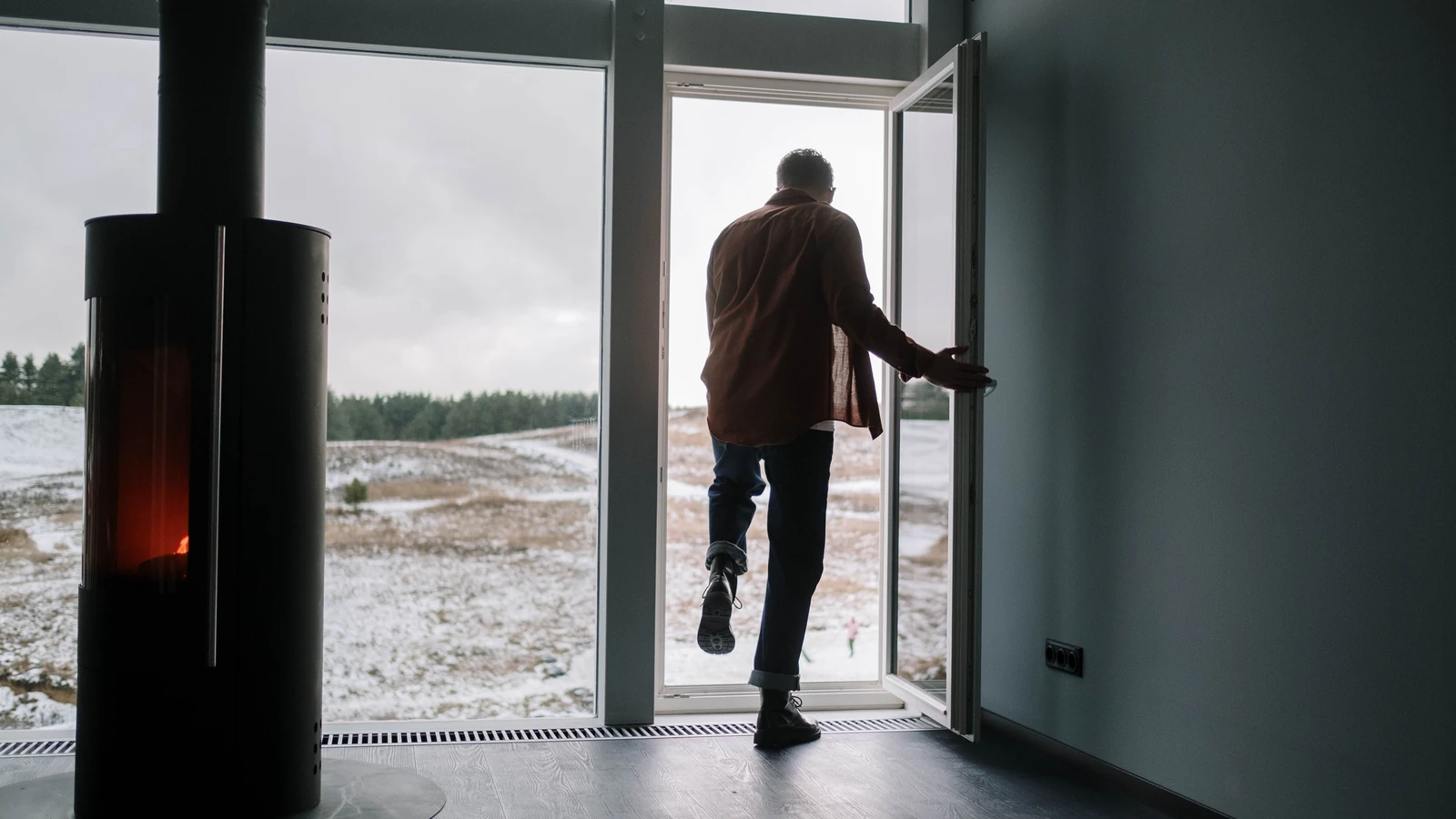 Uomo che esce da una casa moderna verso un paesaggio innevato attraverso una porta di vetro con un camino all'interno.