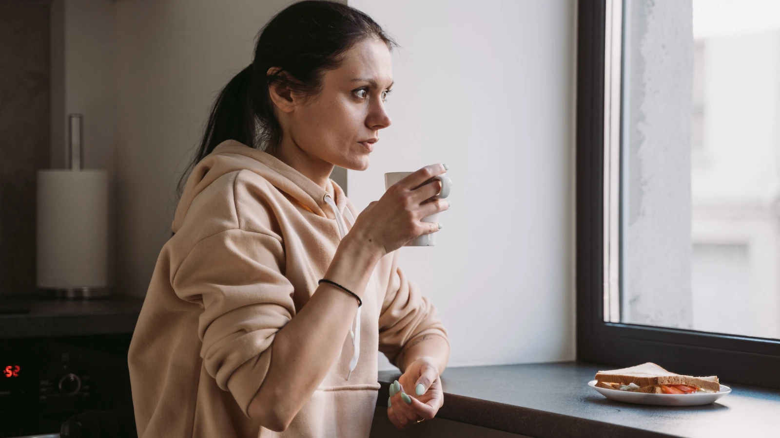 Donna in felpa che gusta una bevanda calda vicino alla finestra con un piatto di cibo accanto. — MART PRODUCTION – Pexels