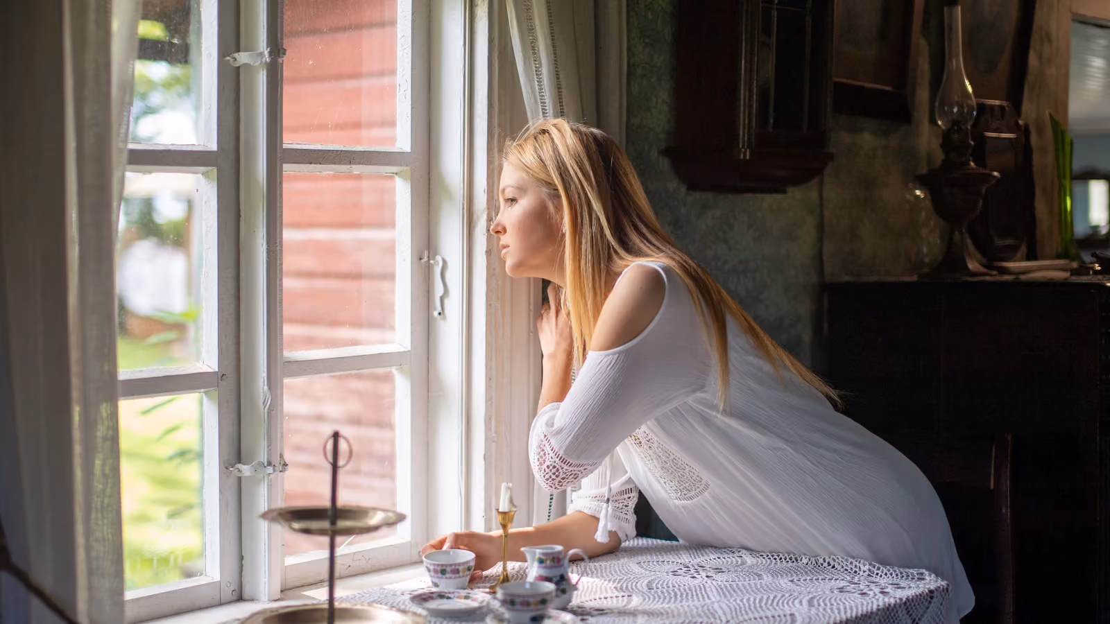 Una donna in abito bianco guarda fuori dalla finestra di un'accogliente stanza rustica a Pringi, in Estonia. — Margo Evardson – Pexels