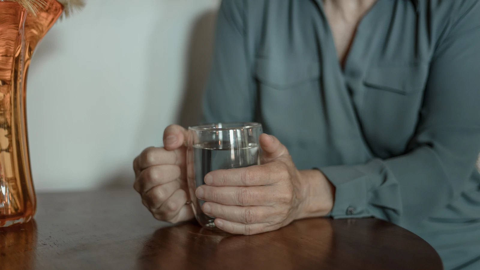 Primo piano di una donna anziana che tiene un bicchiere d'acqua mentre è seduta a un tavolo di legno all'interno.