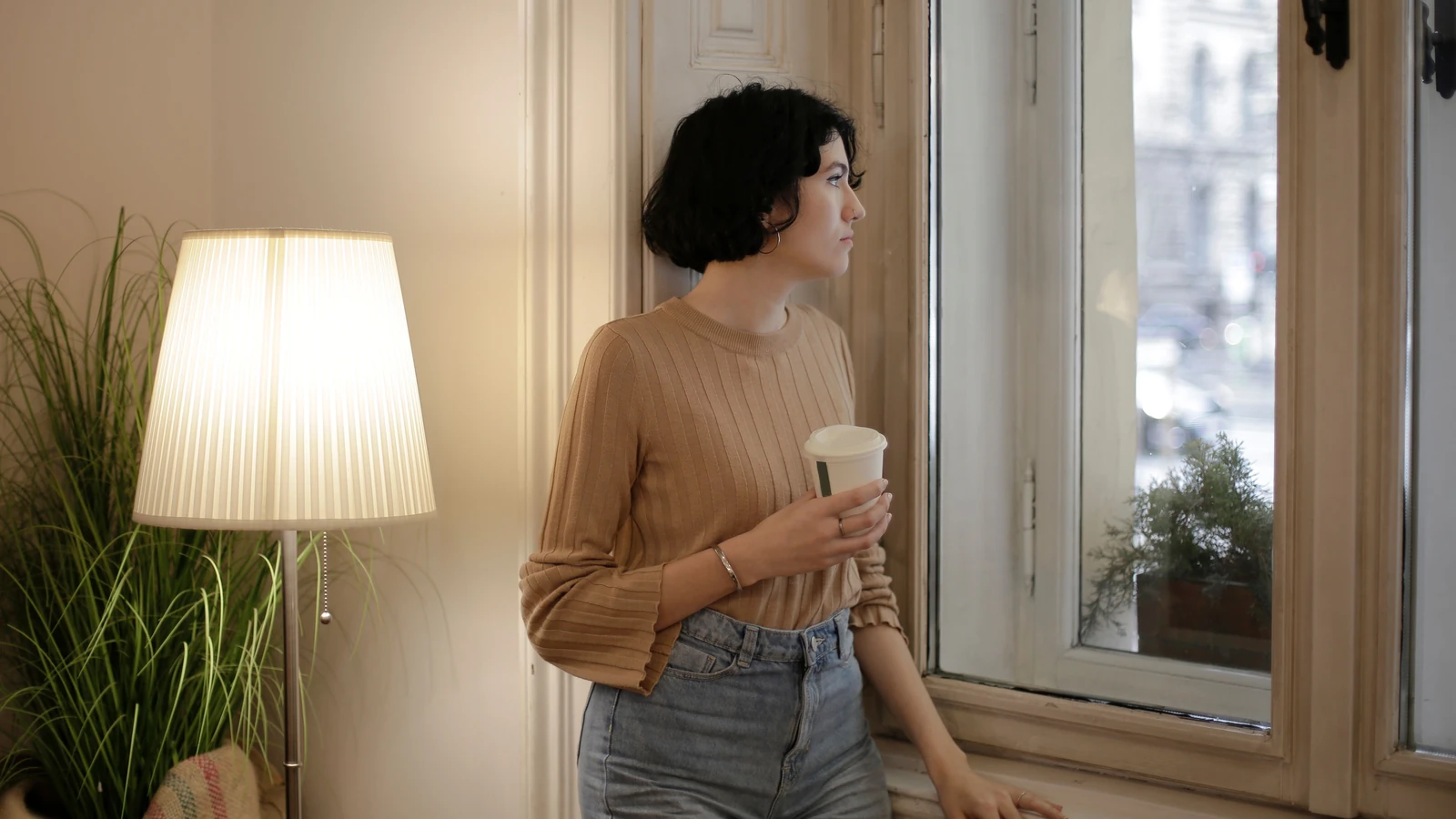 Giovane donna che tiene una tazza di caffè, guarda pensierosa fuori dalla finestra in una stanza accogliente.