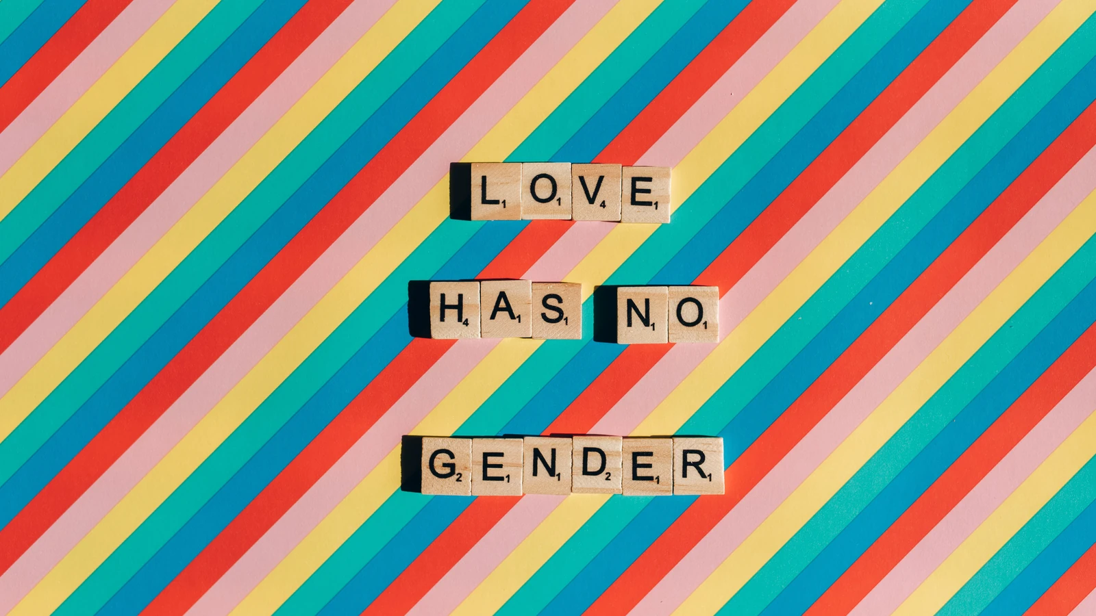 Tessere di Scrabble che formano 'L'amore non ha genere' su uno sfondo a strisce arcobaleno che celebra il Pride.