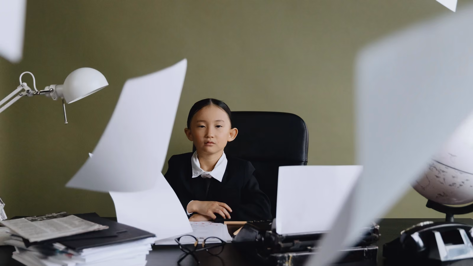 Una bambina in abbigliamento formale è seduta a una scrivania circondata da fogli volanti, che rappresentano creatività e ambizione. — cottonbro studio – Pexels